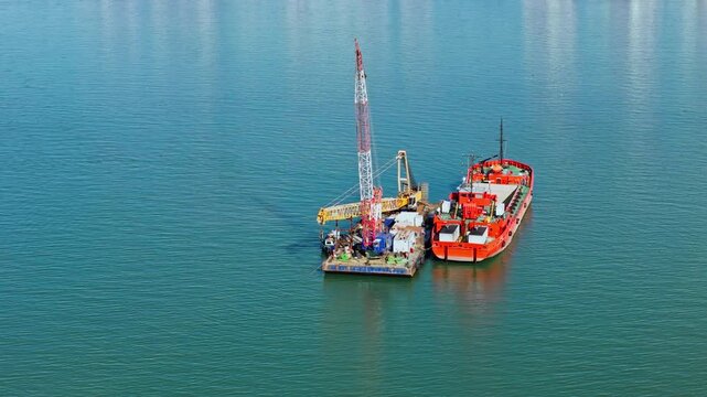 Floating crane platform and dredging vessel during harbor dredging operation. Removal and transfer of seabed material in coastal water