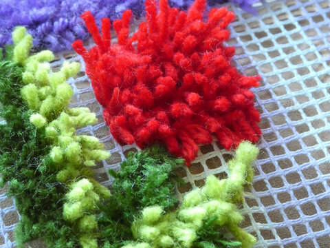 Needlepoint cross-stitch embroidery white grid is plastic canvas or mesh, and the colored fuzzy yarn is being stitched through it to create a raised, textured design, possibly flowers or foliage.