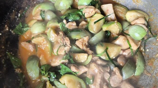 Close-up of simmering Thai spicy stir-fried chicken with green eggplants, basil, and kaffir lime leaves in a wok. 