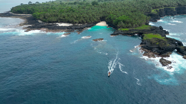 Boat cruising offshore near Ilh&eacute;u das Rolas, S&atilde;o Tom&eacute; and Pr&iacute;ncipe, with Bateria beach and ilheu das rolas  in the background, surrounded by volcanic coastline and turquoise waters.Africa