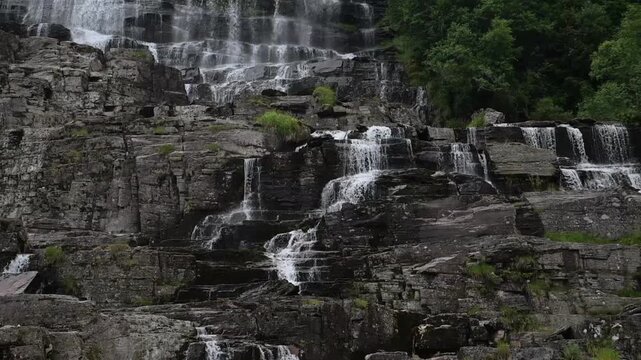 Powerful cascading waterfall flows over dark layered rocks surrounded by lush summer vegetation in Norway.