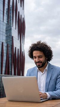 Man with curly hair in blue suit works on laptop outside, symbolizing modern work and urban professionalism