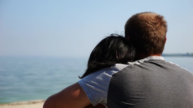Happy pair hugs during sitting on wooden log on beach. Romantic couple admiring sea landscape at sunny day. Lovers spending time together at resort. Concept of summer vacation or holiday. Slow mo