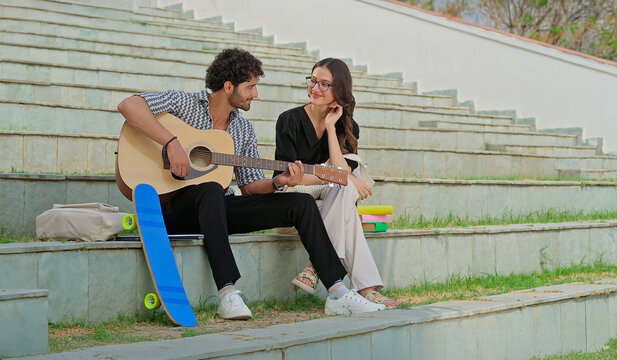 Indian young adult gen z female male man hold guitar play new lyric music sing song have fun joy stairway place. Happy relax teen girl boy guy sitting on stair enjoy good mood spend break day time