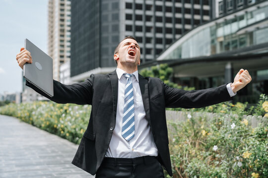 Businessman celebrating success with arms raised holding laptop outdoors, excited professional shouting in joy near modern office building, career achievement concept