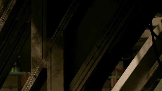 Metal beams and supports are visible in a large factory. The scene shows machinery and tools in the background as workers move around. The lighting is bright and highlights the materials.