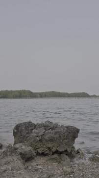 Rocky coastline with volcanic stone and calm ocean water against distant green shore and small house for travel and environmental design context