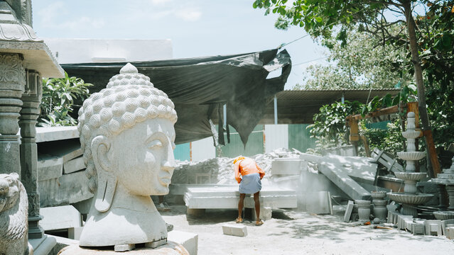 Carved Buddha head sculpture in outdoor stone workshop with artisan working traditional statue craftsmanship