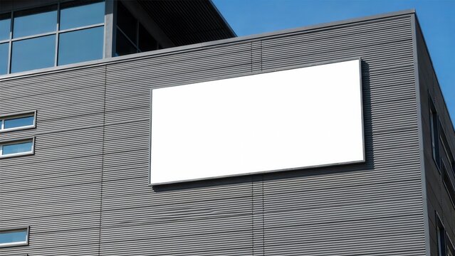 A blank white billboard mounted on a modern dark gray building facade under a clear blue sky. Ideal for outdoor advertising mockups and commercial branding campaigns.