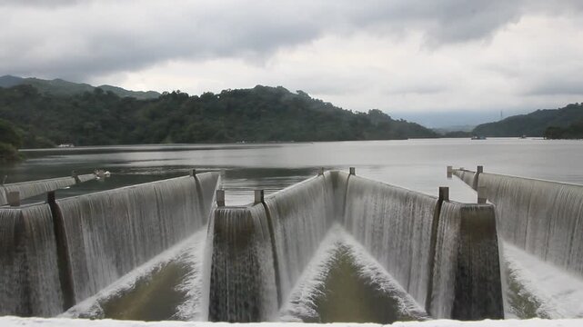 Water overflowing through a labyrinth weir spillway at Liyutan Reservoir in Miaoli, Taiwan. Sawtooth dam spillway controlling water level under cloudy sky.