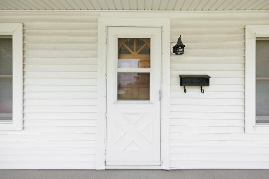 A home's wooden door behind a white screen door with white siding and a black mailbox and light fixture.
