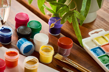 Art supplies and paint containers on a wooden table near a green plant in a well-lit indoor space © mira_y
