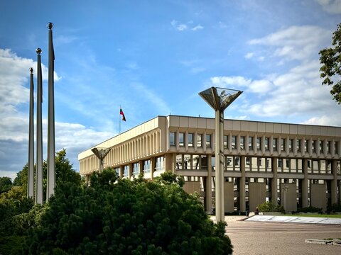 Modernist facade of the Lithuanian Parliament (Seimas) in Vilnius under a blue cloudy sky. Features the national flag, tall flagpoles, green shrubbery, iconic funnel light posts, and paved plaza space