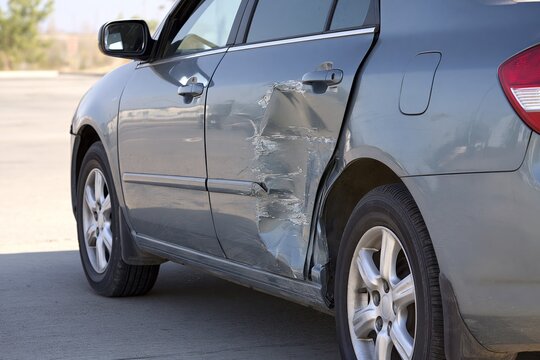 Side view of metallic gray passenger car showing severe collision damage on driver side door with deep dent scraped paint and distorted trim under neutral daylight parked on plain concrete surface