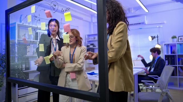 Three businesswoman professionals collaborate on a strategy by writing ideas and notes on a glass board during an office meeting