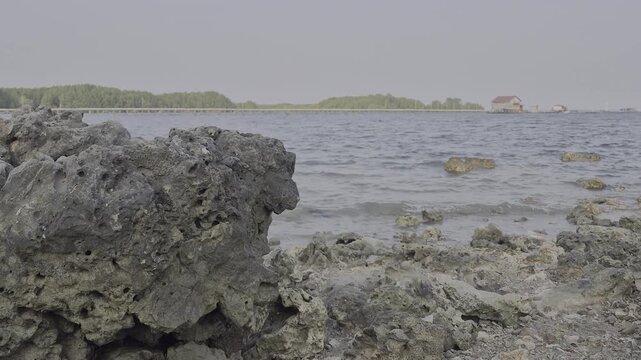 Rocky coastline with volcanic stone and calm ocean water against distant green shore and small house for travel and environmental design context