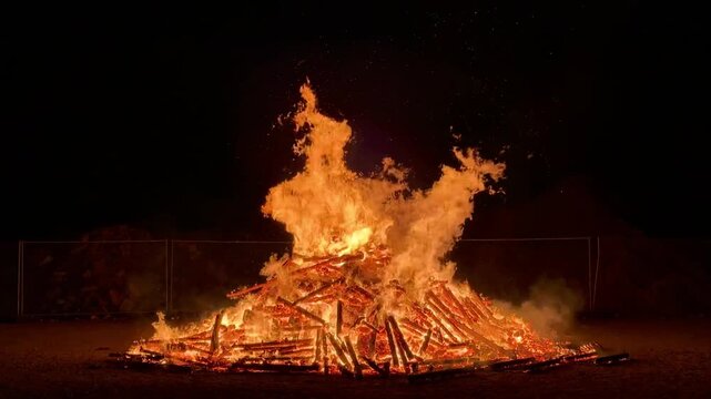 An impressive wide shot of a massive traditional Walpurgis Night (Walpurgisnacht) bonfire made of a giant pile of wood blazing brightly against the dark night sky in Germany.
