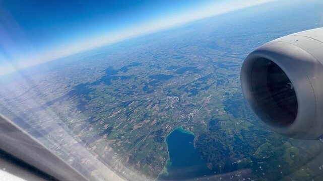 A scenic high-angle video captured through an airplane passenger window, overlooking the picturesque Austrian countryside. The aerial shot reveals neatly divided green agricultural fields, winding