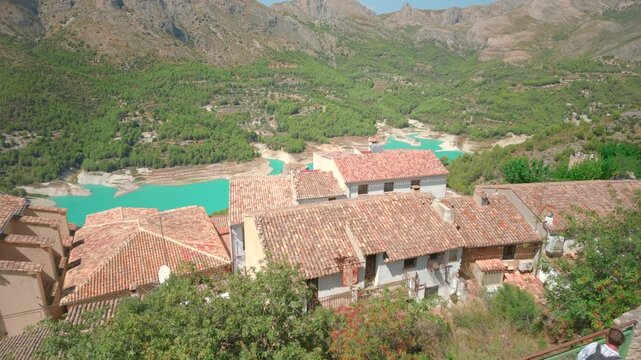 Terracotta roof village above turquoise waterway winding through rocky valley, with green hills, bright daylight, and small visitors on hillside railing overlooking broad scenic panorama