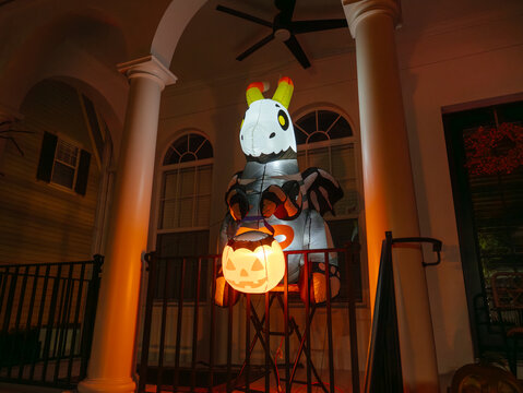 A large inflatable skeleton zebra holds a glowing pumpkin lantern on a decorated house porch. Orange lights illuminate the columns and create a spooky Halloween atmosphere at night.
