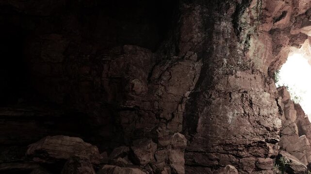 People walk inside a cave that has dark rocky walls. Sunlight shines through a natural opening, creating a contrast with the shadows. The scene shows various rock formations.