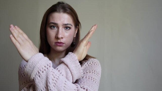 Young adult woman makes a crossed arms stop gesture while looking seriously at camera expressing refusal rejection prohibition boundaries and a clear message to say no