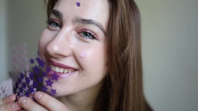 Vertical screen smiling young woman holds purple star shaped acne patches by her face while wearing stickers showing playful skincare and targeted care for skin blemishes