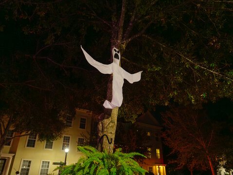 A white ghost decoration hangs from a large tree in a residential neighborhood. Night lighting creates a spooky Halloween scene among dark branches and houses.