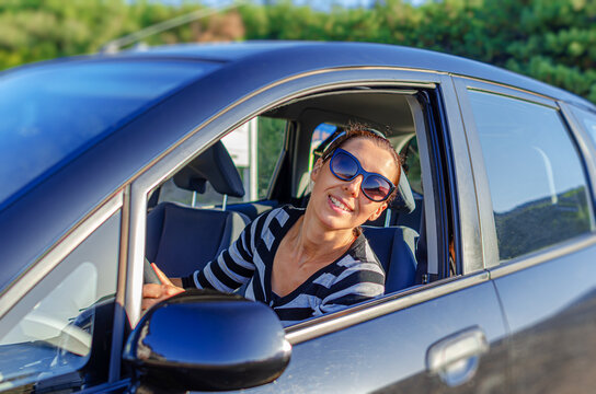 A happy woman wearing sunglasses smiles out of an open car window on a bright sunny day.