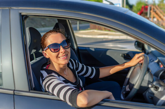 A smiling woman wearing sunglasses sits in the driver's seat of a black car with her hand on the steering wheel during a sunny day.