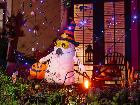 A white inflatable ghost wearing a striped witch hat stands beside a pumpkin on a residential porch. Purple lights, spiders, webs, and eyeballs create a colorful Halloween display at night.