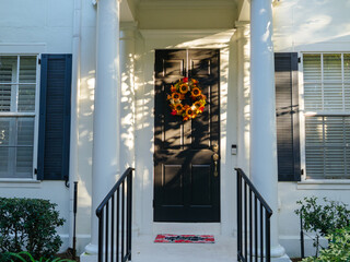 Naklejka premium A welcoming home entrance features a colorful autumn wreath on a black front door framed by elegant white columns. Soft afternoon light creates a warm seasonal atmosphere.