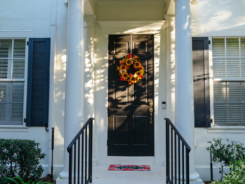 A welcoming home entrance features a colorful autumn wreath on a black front door framed by elegant white columns. Soft afternoon light creates a warm seasonal atmosphere.