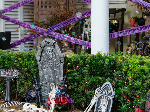 A Halloween yard display features tombstones, skeleton bones, colorful flowers, and haunted caution tape. Floating bubbles add an unexpected playful touch to the spooky outdoor scene.