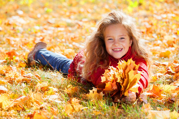 Naklejka premium Little girl lying on grass covered with autumn leaf holding yellow maple leaf bouquet and smiling. Happy childhood outdoor play in fall park. Joy and nature concept