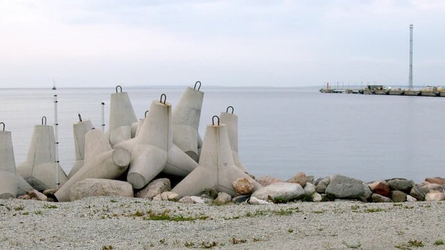 Wide static 4K view of concrete wave breakers and rocks protecting the shoreline in Tallinn, Estonia, with calm Baltic Sea water and harbor infrastructure in the distance under an overcast sky