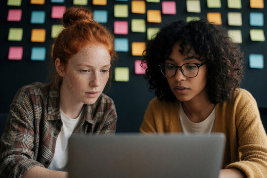 Two young women focus on laptop screen. Colorful sticky notes adorn wall behind them, indicating creative workspace. They engage in collaborative project, sharing ideas and concentrating on tasks.