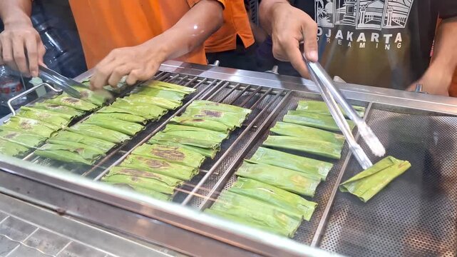 Hands turn leaf-wrapped food over a hot grill. Steam rises as food cooks evenly on a metal grate. Busy street vendor prepares popular local delicacy. Vibrant green leaves show signs of grilling