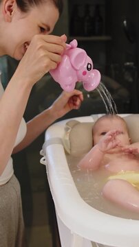 Mother gently pours water on baby during bath time using a pink elephant toy for playful splashing. The scene captures playful interaction and a warm family bonding moment.