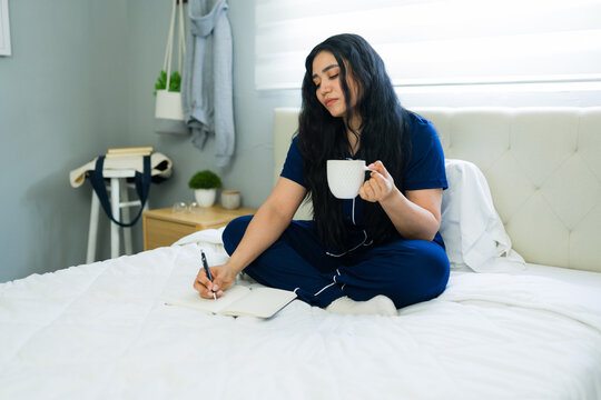 Serious woman writing in a journal and drinking coffee in bed, enjoying a relaxing morning routine at home