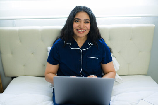 Happy woman wearing pajamas, sitting in bed, smiling and typing on a laptop, enjoying a comfortable remote work morning