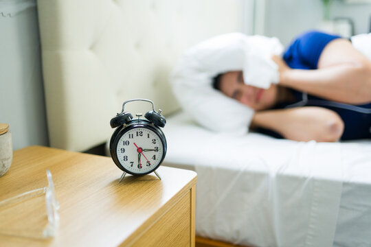 Woman trying to sleep, covering head with pillow, disturbed by loud alarm clock ringing early in morning