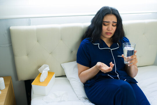 Young woman feeling unwell, taking medicine with water, sitting on bed at home during an illness