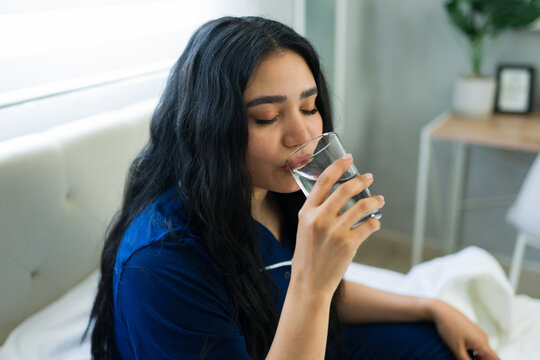 Healthy woman in pajamas drinking water while sitting on bed, practicing good hydration habits in the morning