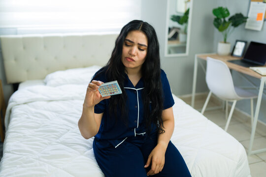 Hispanic woman in pajamas sitting on her bed, looking concerned while holding a pack of oral contraceptive pills