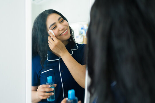Woman in pajamas cleaning her face with micellar water and cotton pad, removing makeup for nightly skincare routine