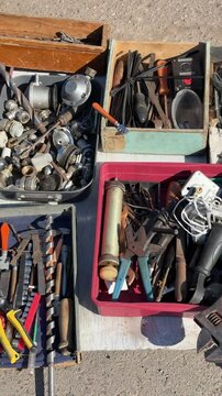 Used tools displayed on asphalt, assorted secondhand hand tools and small parts spread across open toolbox and plastic trays