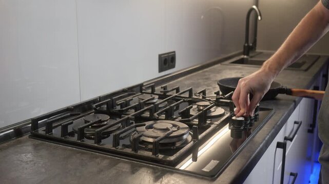 Hand places frying pan onto gas cooktop in modern kitchen at low oblique angle, with pan, fryingpan, cooktop, stovetop detail, showing gas, grate, burner, knob context