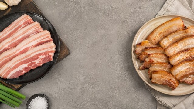 Raw and grilled pork belly slices on plates. Korean BBQ samgyeopsal dish with garlic, green onion and salt on gray stone table. Asian cuisine cooking concept with copy space.