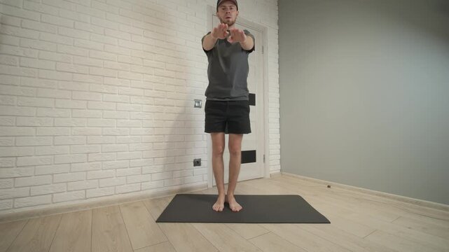Male subject performs low squat then returns to upright standing posture on black exercise mat inside small home workout area, with man, squat, stand, exercise detail, showing workout, fitness, mat